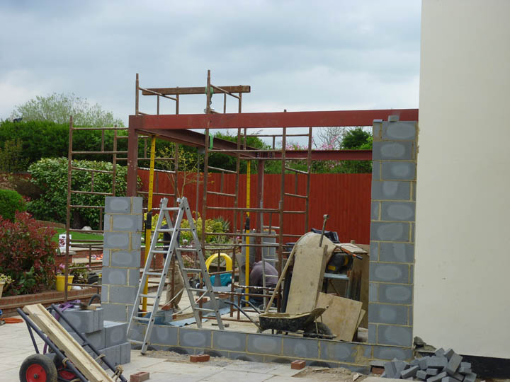 Orangery Build Project Portal Frame. This orangery will feature large hardwood folding-sliding doors and narrow masonry pillars, therefore we need to use a steel portal frame to strengthen the structure and give the building rigidity. The vertical columns are bolted to the foundations using resin-bonded steel studs. These columns are connected to the steel ring-beam and tied back to the main house. There are brick-ties welded to the post to connect the blocks. The guys have used a hoist arrangement to lift the steel beams into place, and support props to correctly position them while they are bolted and welded together. The adapted carpet trolley in the foreground of the photo is one of the best items of equipment we've ever invested in; it make handling large steel and glulam beams, simpler and safer.