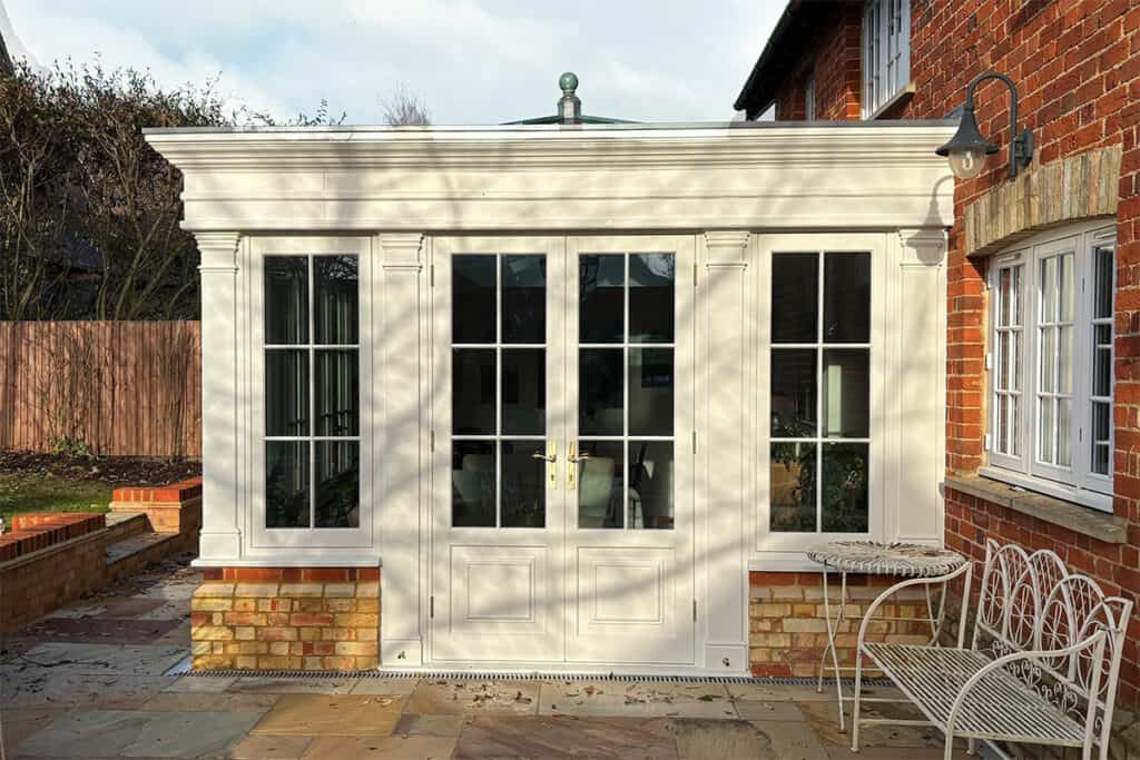 Cream-painted timber orangery with French doors, decorative cornice and brick base